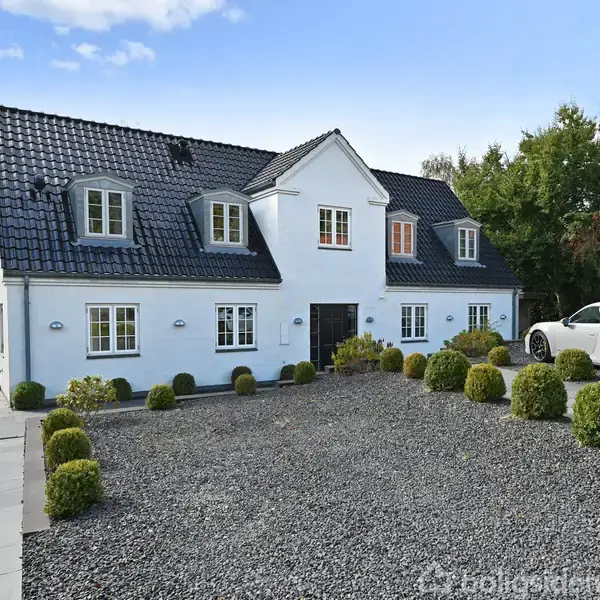 En hvid to-etagers villa med sort tag står foran en grusbelagt gårdsplads omgivet af runde buske; biler er parkeret til højre, og et grønt område ses i baggrunden.