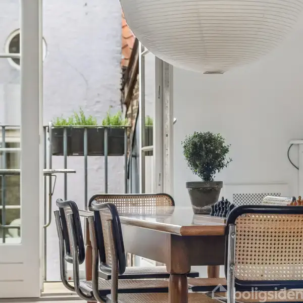 Et spisebord med fire stole står i et lyst rum. En stor rund lampe hænger over bordet, og en potteplante er placeret midt på bordet. En fransk altandør er åben i baggrunden.