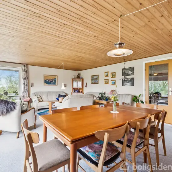 Trælakeret spisebord med seks stole står i en stue med lyst træloft. Sofasæt og dekorative billeder pryder væggene. Store vinduer giver udsigt til en grøn have.