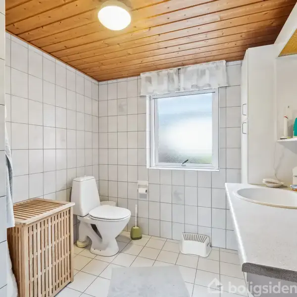 Badeværelse med toilet, håndvask og opbevaringsskab. En vasketøjskurv står ved siden af toilettet. Rummet har hvide fliser, et træloft og et vindue med let indfald af dagslys.