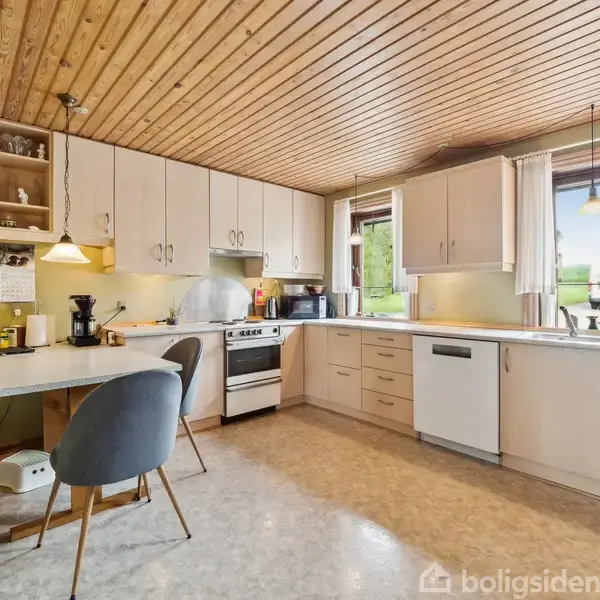 Et køkken med hvide skabe og grå bordplader. En ovn og opvaskemaskine står indbygget. Der er et lille spisebord med stole samt store vinduer med udsigt til en grøn have.