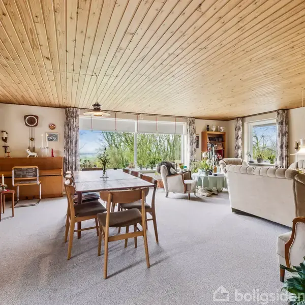 Stue med trægulv og store vinduer, indeholder spisebord og stole i midten. Sofa og lænestole er placeret ved vinduerne. Indretningen er retro, med træmøbler og planter.
