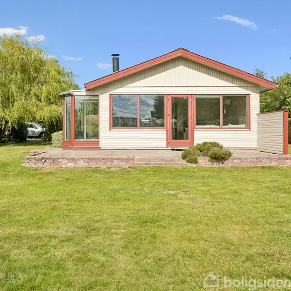 Et lille, hvidt hus med røde detaljer står i en grøn have. Store vinduer reflekterer solen. En grussti leder til hovedindgangen. Træer omkranser ejendommen i en rolig landskab.
