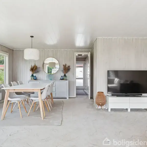 Spisestue med hvidt bord og seks stole, placeret ved store vinduer. Et spejl og dekorationer på en skænk. TV på hvidt møbel til højre. Lys indretning.