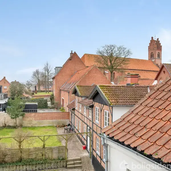 Røde teglstenstage dækker smukke murstenshuse, træer i baggrunden. Kirketårn rejser sig mod en blå himmel i en rolig bydel, omgivet af grønne haver og hække.