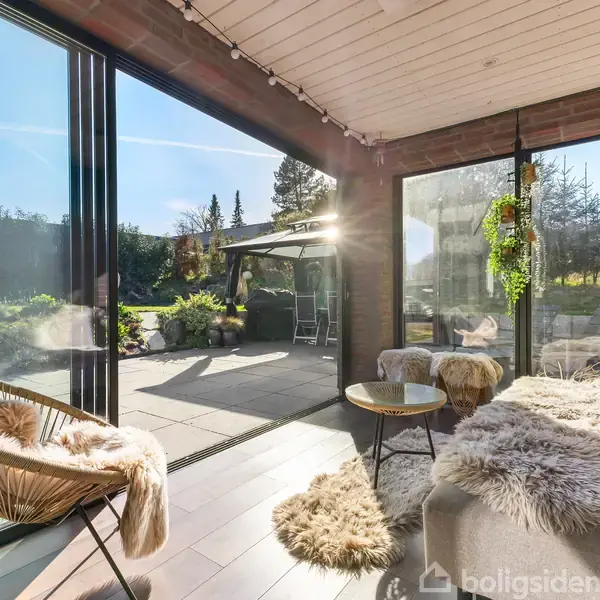 En lukket terrasse indeholder komfortable stole og en sofa med pelsbetræk. Sollys skinner ind gennem store vinduer, der giver udsigt til en udendørs terrasse med planter og klart himmel.