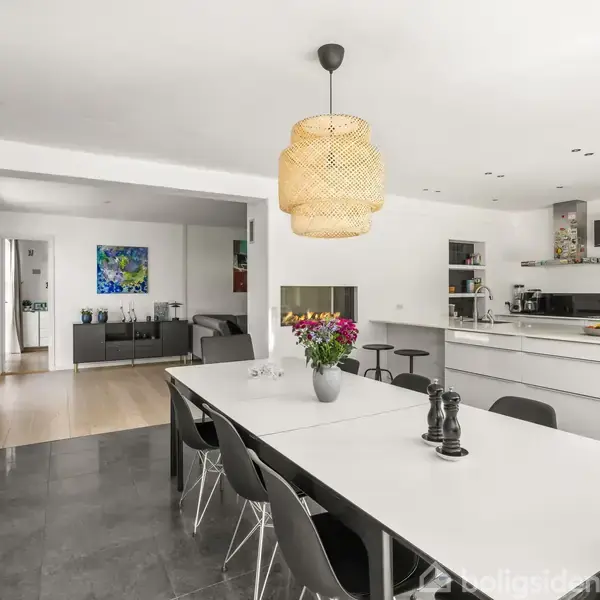 Spisebord med sorte stole står i et lyst køkkenalrum. En pendellampe hænger over bordet. I baggrunden ses et moderne køkken med en ø og et stueområde.