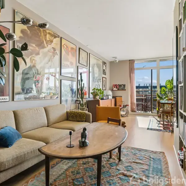 Stue med beige sofa og sofabord, dekoreret med plakater. Rummets baggrund viser en balkon gennem glasdøre, med udsigt til byen. Flere planter skaber en grøn atmosfære.