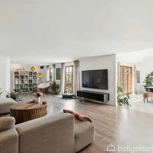 Stue med en beige sofa, et rundt træbord, og en stor fladskærm på væggen. I baggrunden er der en bogreol og et spiseområde med glasdøre til haven.