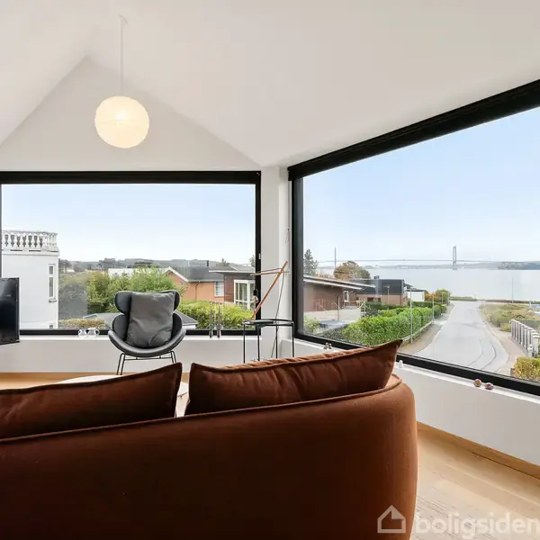 Stue med brun sofa og lænestol, der vender mod store vinduer. Udsigt over et boligområde og en fjord i baggrunden. TV i hjørnet, moderne indretning.