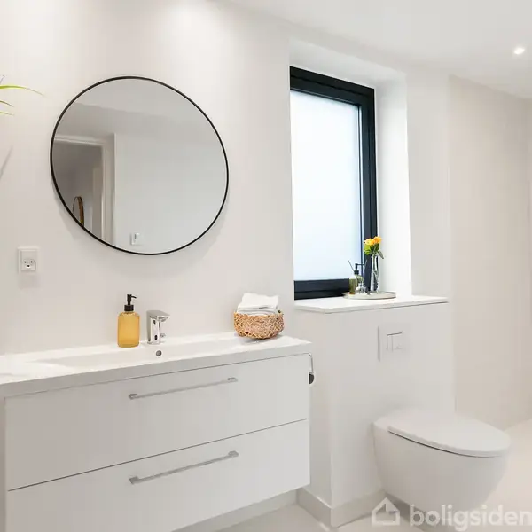 Et moderne badeværelse med hvide møbler og vægge. Rundt spejl over en vask med sæbe og en lille kurv. Bruser ved siden af vinduet med en blomstervase.