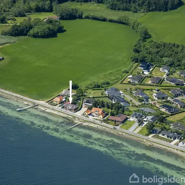 Kystnært boligområde med huse beliggende ved vandet og marker i baggrunden. En pil peger på et specifikt hus nær stranden. Grønne områder omgiver nabolaget.