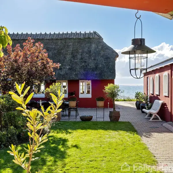Rødt hus med stråtag står foran en terrasse, sand og havudsigt. Grønne planter omgiver gårdspladsen med bord og stole. Hængebriks lyser fra oven.