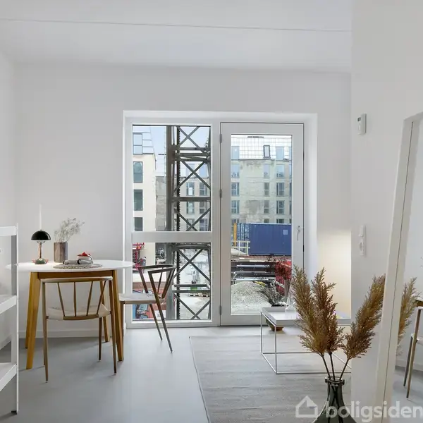 Et lyst rum med et skrivebord, stol og hylder til venstre. En stor glasdør åbner til en altan med udsigt over bygningskonstruktioner. Spejl og dekorationer til højre.