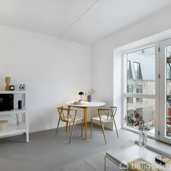 To stole og et rundt bord står ved et stort vindue i en minimalistisk, hvidmalet stue. Et hvidt reolsystem med mikroovn står til venstre. Ingen tekst.