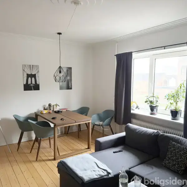 En stue indeholder et skrivebord med to stole, omgivet af malerier på væggen. Ved vinduet står planter. Foran skrivebordet er en grå sofa ved et stort vindue med mørke gardiner.