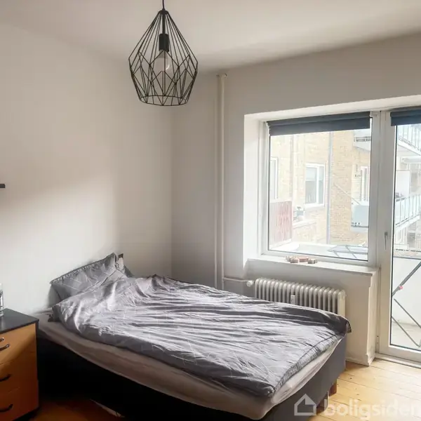 Et soveværelse med en seng der har grå sengetøj, står ved siden af et vindue. En lampe hænger fra loftet; der er en radiator under vinduet.