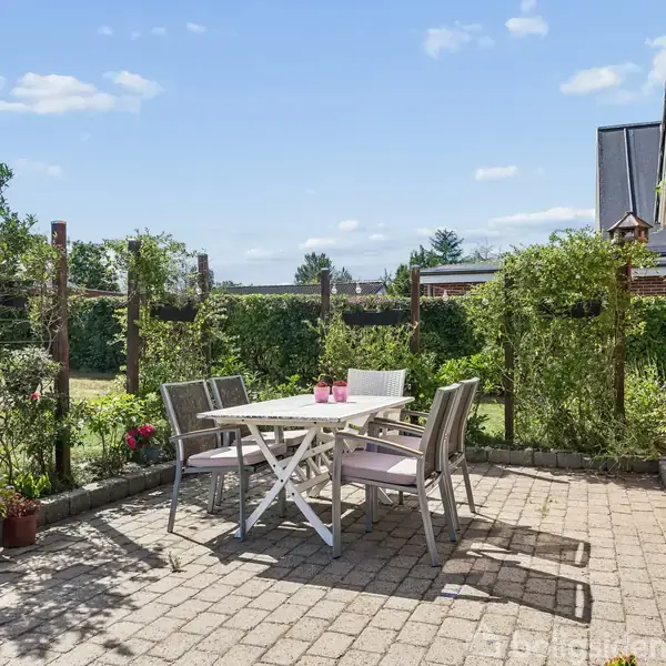 Terrassemøbler står på flisebelagt område med bord og fire stole. De omgives af grønne buske og blomster, og en murstensvæg ses til højre under en klar blå himmel.