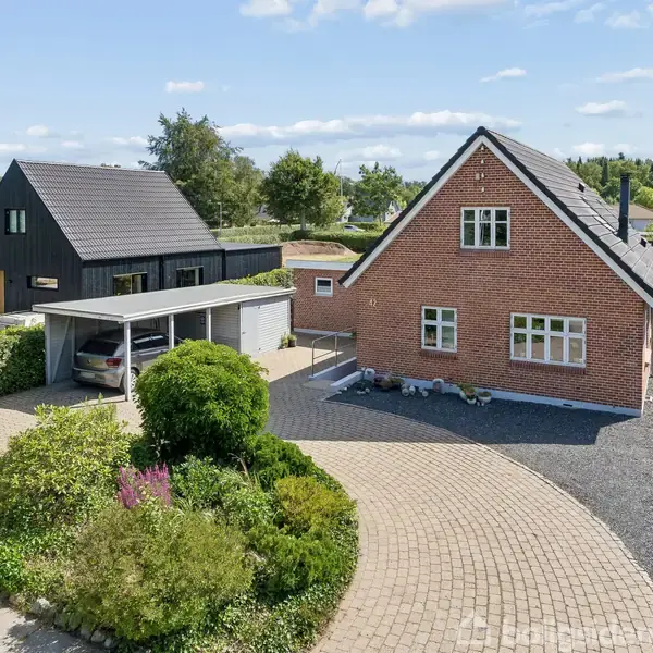 Rød murstenshus med sort tag står stille ved siden af en carport med en bil. Omkring er der en velplejet have med buske og en brostensbelagt indkørsel. Tegn med teksten "Til salg".