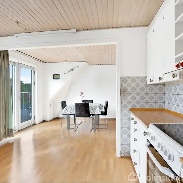 Køkken med hvidt interiør, åben mod stue med spisebord og fire stole. Store vinduer med grønne gardiner giver udsigt til altan og omgivelser. Lyst trægulv og mønstrede fliser.