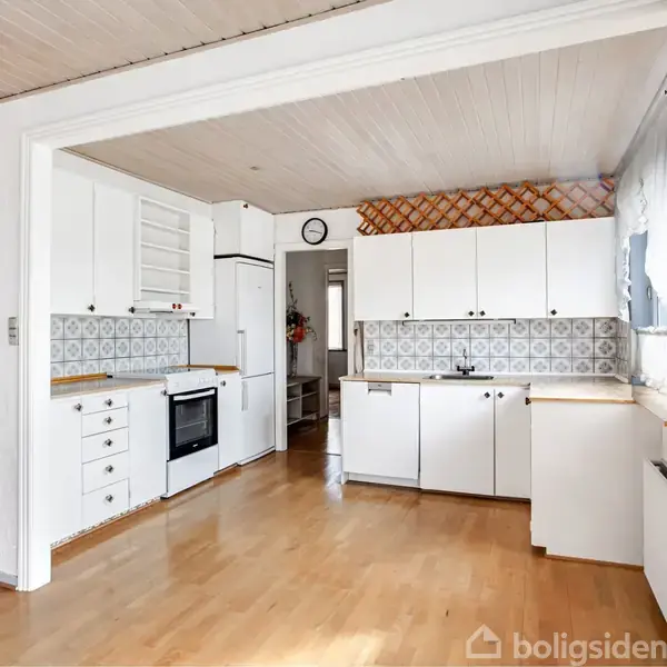 Hvidt køkken med træskuffer og skabe, placeret langs væggene. Ovn og kogeplader er indbygget. Mønstret flisebagvæg. Trægulv og stort vindue med gardiner. Vægur hænger på væggen.