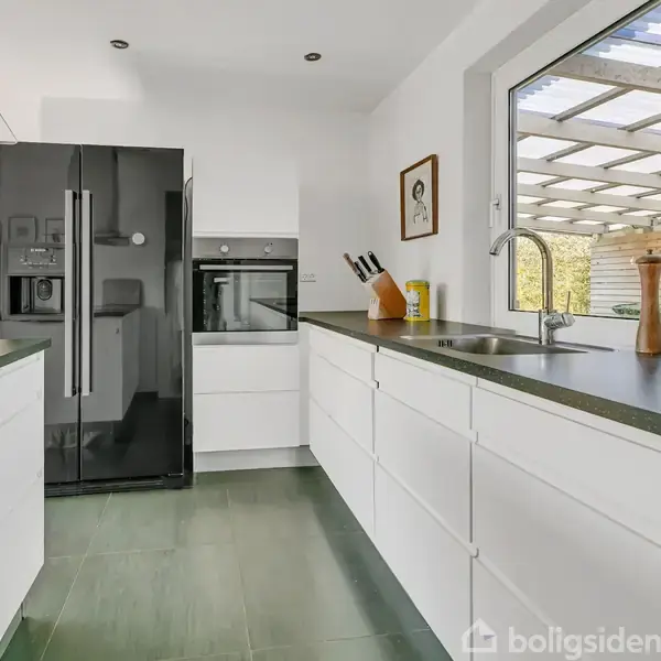 Et moderne køkken har en sort køleskab, hvide skabe og en vask. En knivblok og peberkværn står ved vinduet, som åbner ud mod en terrasse med pergola.