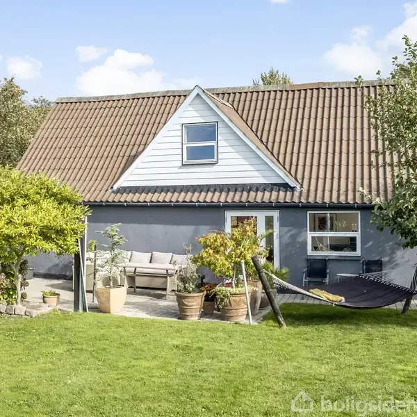 Et hus med rødt tegltag og grå vægge står i en have. En solbeskinnet terrasse har havemøbler og en hængekøje omgivet af grønne træer og buske.