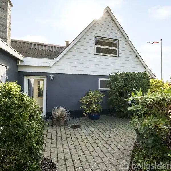 Et gråt hus med en trekantet gavl står stille omkranset af grønne buske. En flisebelagt sti fører til hoveddøren. Et flag vajer i baggrunden.