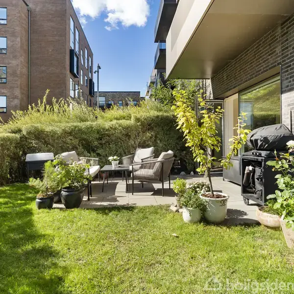 Terrasse med havemøbler og grill står på en flisebelagt overflade. Omgivet af grønne planter og tæt klippet hæk. Moderne lejlighedsbygninger ses i baggrunden under en klar blå himmel.