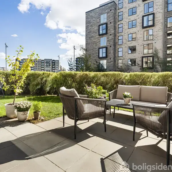 Terrassemøbler står på flisebelagt terrasse, omgivet af grønne planter og buske. I baggrunden ses en høj, moderne beboelsesejendom under en solrig, blå himmel.
