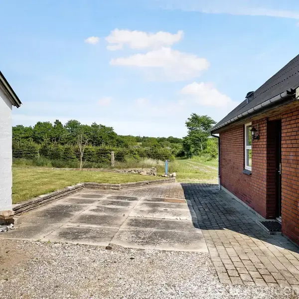 En rød murstensbygning med et tilstødende indkørselsområde af betonfliser. Til venstre ses en hvid bygning med en grøn garageport. Baggrund præget af græsplæne og træer under en blå himmel.