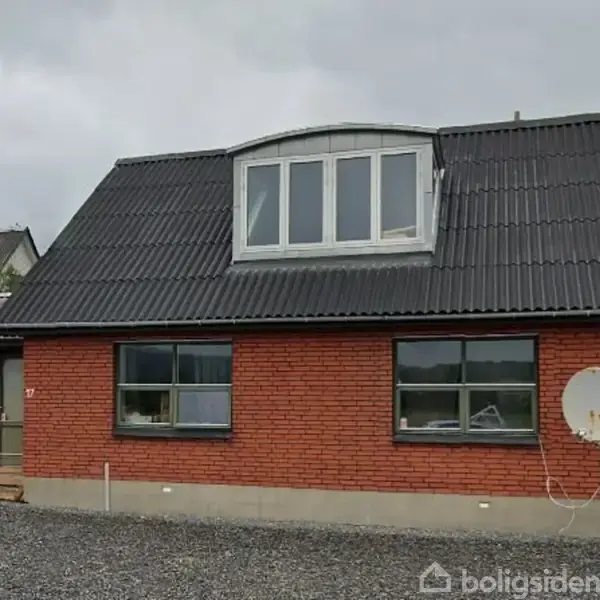 Murstenshus med sort tag, vinduer på første sal, satellitparabol på væggen. Grusbelagt forhave og en skyet himmel i baggrunden.