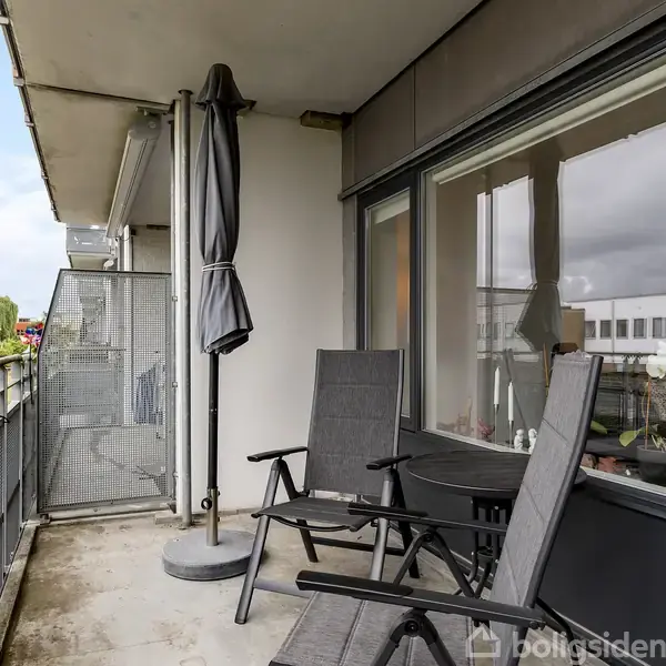 Sort lænestol med fodskammel står på et lyst trægulv ved siden af et sofabord og en grå sofa. Rummet har en hvid væg, gulvlampe og planter.