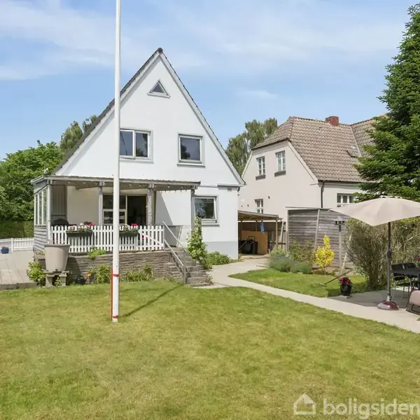 En hvid villa med trekantet gavl står i en velplejet have med græsplæne. Foran huset er en terrasse med havemøbler og parasol.