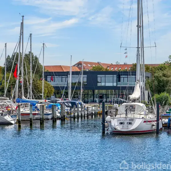 Både ligger til kaj i en marina, hvor de roligt flyder på vandet. I baggrunden ses en moderne bygning og træer, der skaber en hyggelig havnestemning.