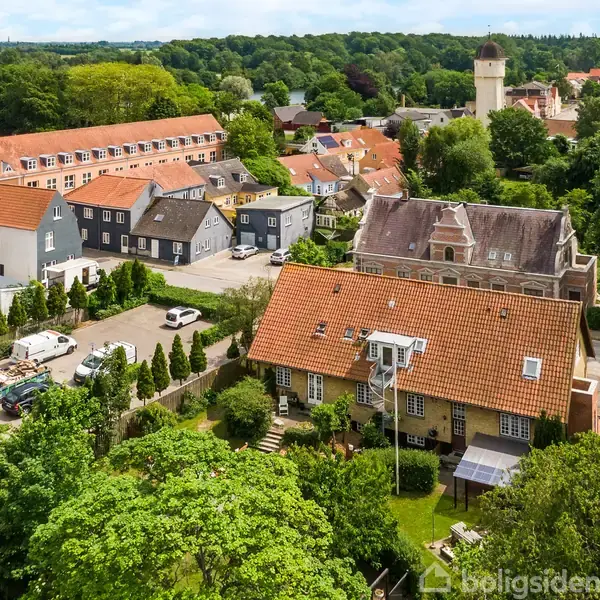 Rødt tegltagshus, omkranset af træer og grønne haver, står stille midt i en dansk landsby. Mange huse og en kirke med tårn ses i baggrunden under en blå himmel.