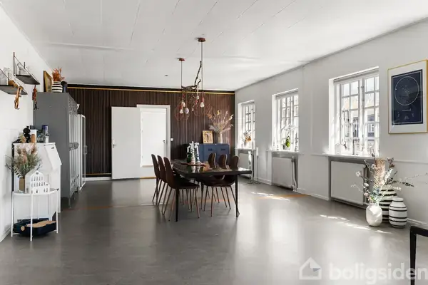 Spisebord med stole i store, lyse spisestue. Hængende lamper over bordet, dekorerede vægge med billeder. Vinduer langs højre side, stor gulvplads, minimalistisk indretning med moderne detaljer.