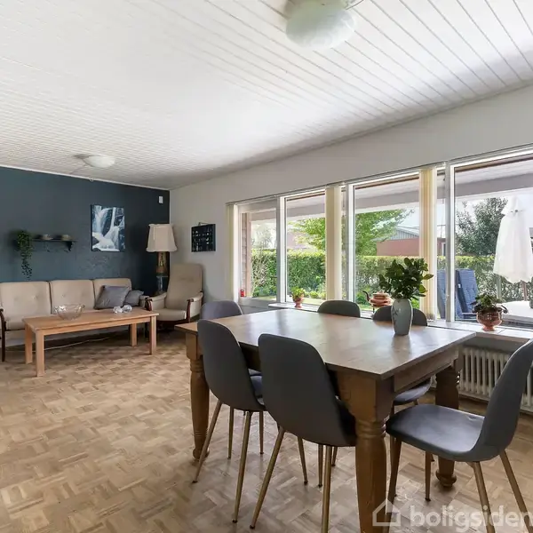 Stue med spisebord og seks stole foran store vinduer, der giver udsigt til terrasse og have. Sofaarrangement med sofabord og lamper i baggrunden. Lyse træbjælker i loftet.