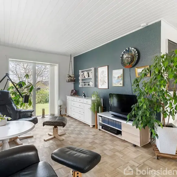 En stue med sorte lænestole og fodskammel står nær et rundt sofabord. Mod en mørk væg er der en kommode, hylde, tv og planter. Store vinduer viser en have.