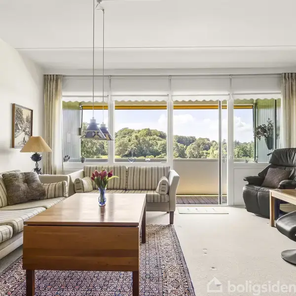 Stue med stribede sofaer, et sofabord med blomster og lænestole, foran store vinduer med udsigt til grøn landskab og altan. Væggene har kunstværker.
