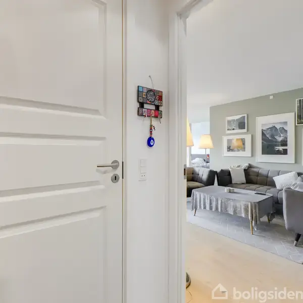 Dør åbner ind til en stue med en grå sofa og sofabord på gulvtæppe. Væggene har flere indrammede billeder, og en gulvlampe står ved sofaen.