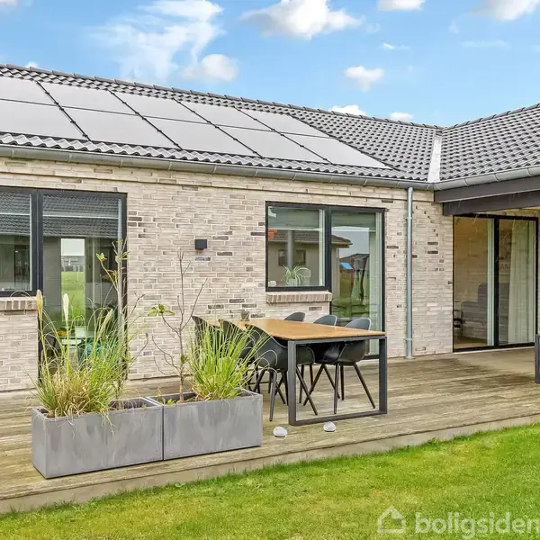 Et moderne hus med solpaneler på taget. En træterrasse har et bord og stole, omkranset af grønne planter. Græsplæne strækker sig foran terrassen under en blå og skyet himmel.