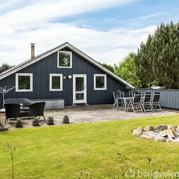 Et sortmalet sommerhus står stille på en græsplæne. Foran ses en møbleret terrasse med borde og stole. Rundt omkring er der grønne buske og træer.