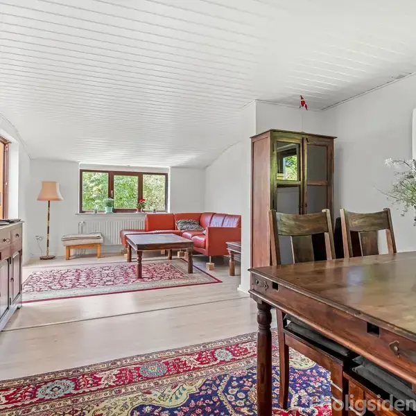 Stue med træmøbler, hvor et spisebord står i forgrunden og en rød sofa ved et sofabord i baggrunden. Store vinduer lukker lys ind, og tæpper dækker gulvet.