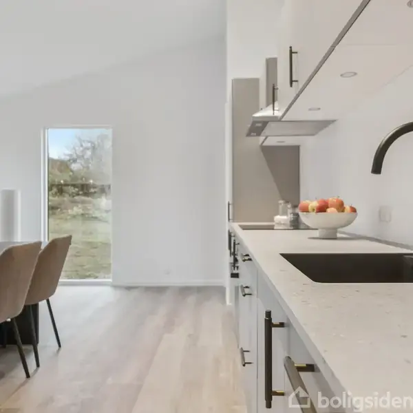Køkkenområde med hvid bordplade og sort vask i forgrunden, frugtskål ved siden af; spisebord med seks stole i baggrunden. Rummet er lyst med udsigt til have gennem glasdør.