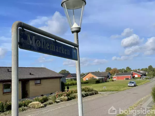 Et vejskilt med teksten "Møllemarken" står ved en brolagt vej. Omgivelserne består af huse med haver og en blå himmel med få skyer.