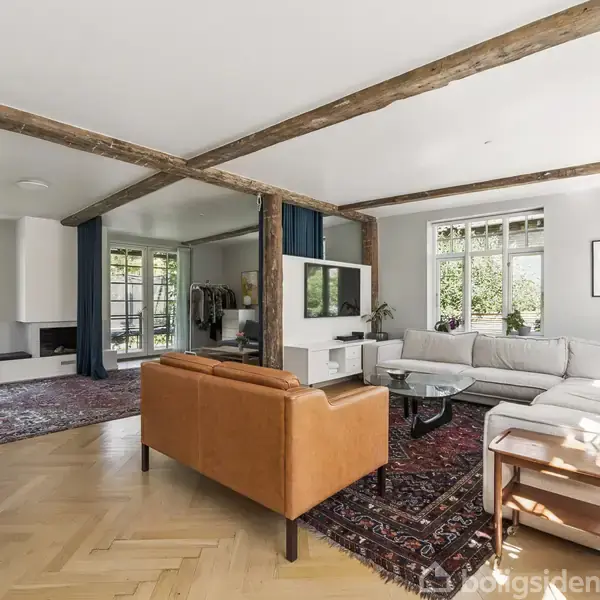 Et moderne stueområde med hvide vægge, træloftsbjælker og store vinduer. En brun lædersofa og en lys hjørnesofa står på et persisk tæppe. Grønne planter og en væglampe skaber hygge.