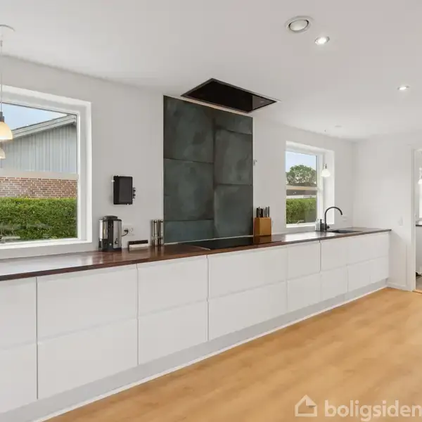 Køkken med hvide skabe og mørk bordplade, let belysning fra to vinduer, udsigt til en hæk og murstensbygning udenfor. Hængelampe og skærebræt til højre.