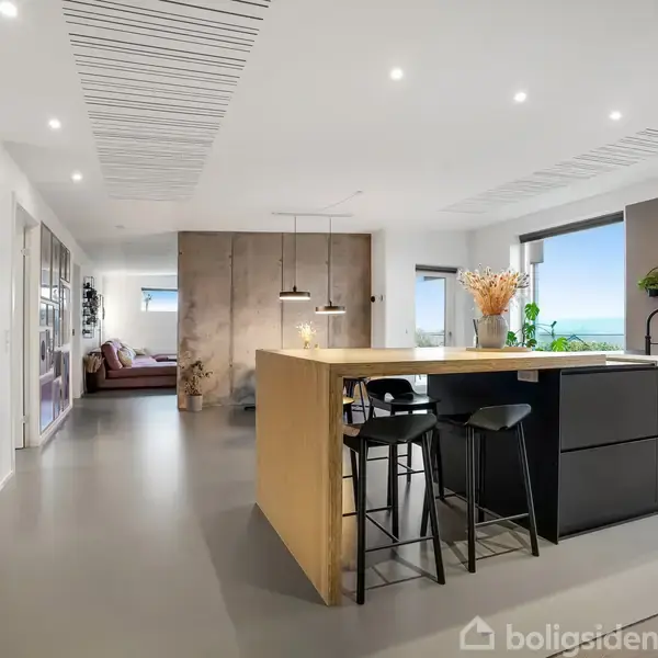 Stue/køkken-område med moderne indretning, hvor en stor køkkenø med barstole dominerer midten. Rummets store vinduer giver udsigt til landskabet og belyser det minimalistiske interiør.