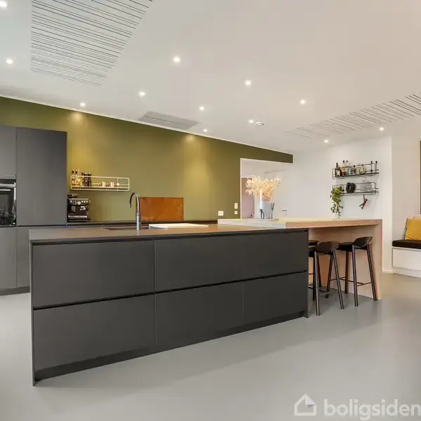 Køkken med sort køkkenø og integreret ovn, rummelige skuffer. Grønmalt væg med hylder, barområde med tre høje stole. Hyggelig siddekrog med puder og dekorative elementer. Moderne belysning.